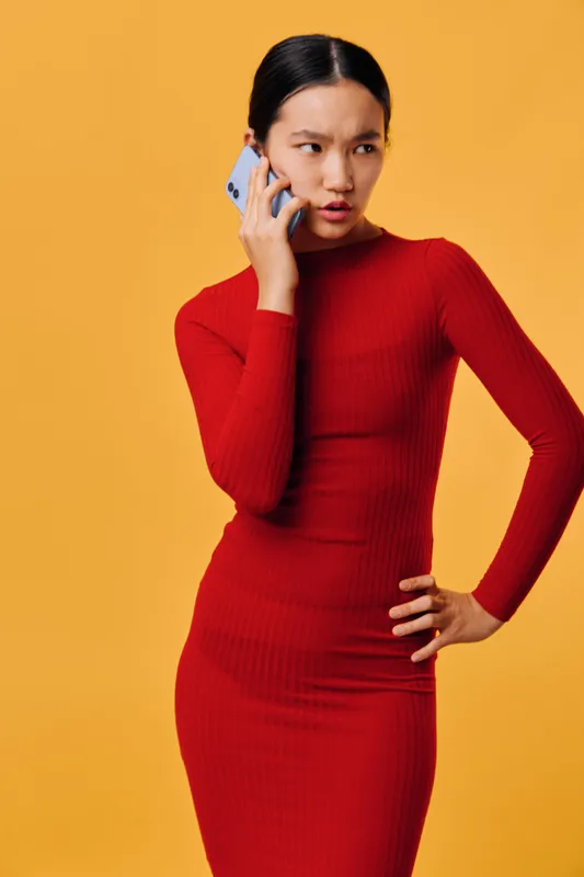 Woman with smartphone wearing red dress talking on phone with thoughtful expression on yellow background. Portrait of young female communication technology concept and mobile call.