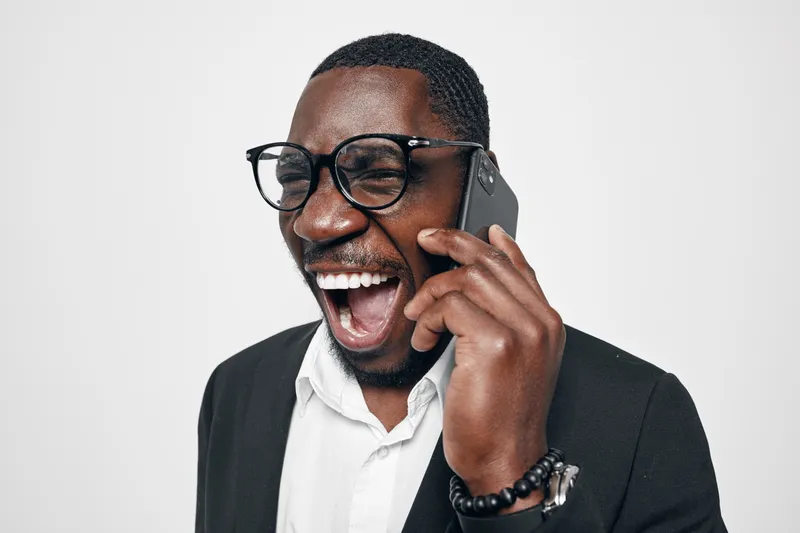 African American man with dark skin wearing glasses and black suit talking on smartphone laughing with joy isolated on white background. Happy male professional in business attire.