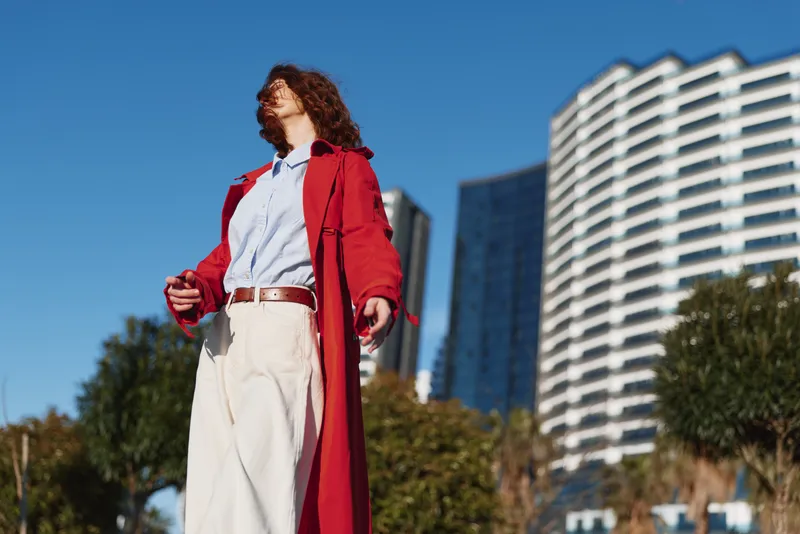 Young woman in a red coat confidently walks outdoors in an urban setting with modern buildings under clear blue sky, expressing freedom and confidence.