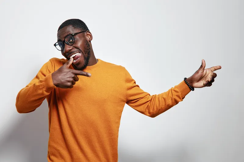 African American man with dark skin wearing glasses and orange sweater smiling and pointing fingers in playful gesture on white background.