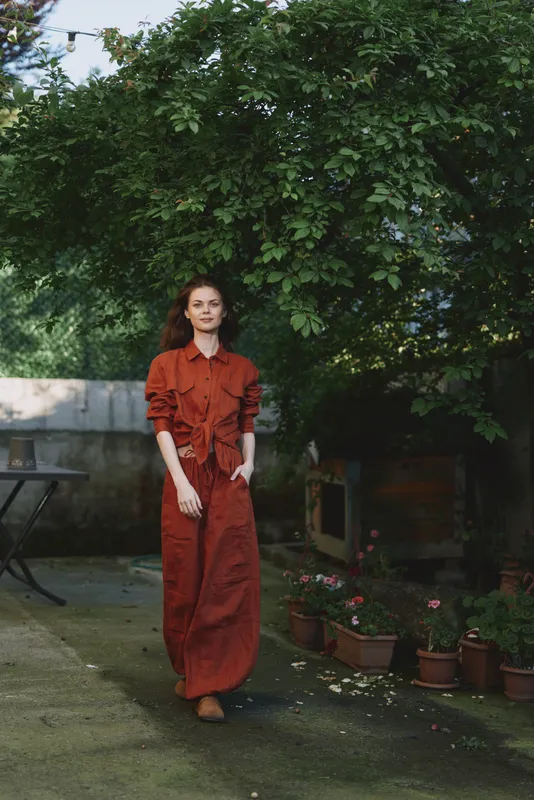 A young woman in a rust red dress strolls through a sunlit garden, her calm gaze framed by lush greenery as terracotta pots line the path