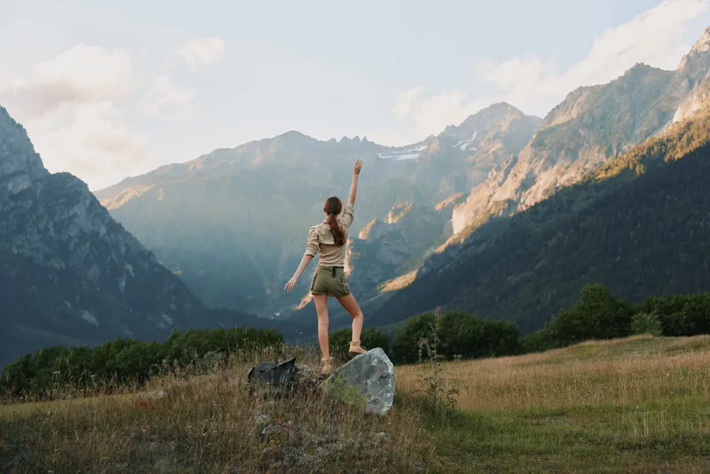A young woman with long hair stands on a rock in a mountain meadow raising her arm, enjoying the natural landscape and freedom of the outdoors.