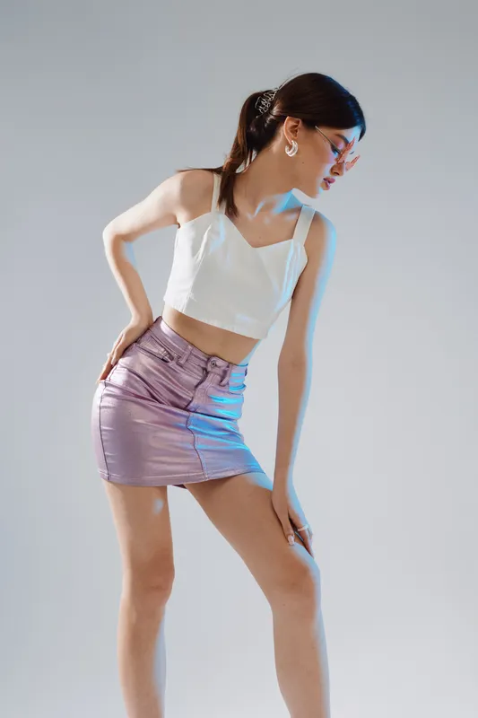 Young woman in a white top and purple skirt poses fashionably against a light background. She wears glasses and earrings, showcasing beauty and modern style.