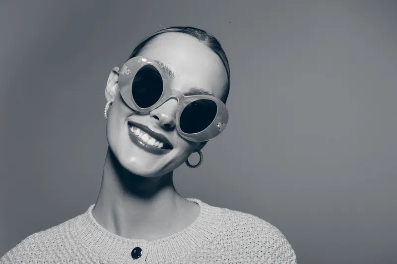 Smiling white woman with pale skin wearing round sunglasses and knitted sweater poses in studio, showing joyful mood and modern fashion style.