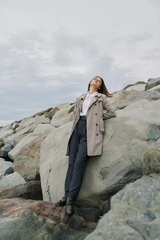 Young woman in casual coat leans against large rocks outdoors, eyes closed and head tilted back. Cloudy sky background creates calm mood and sense of freedom and relaxation.