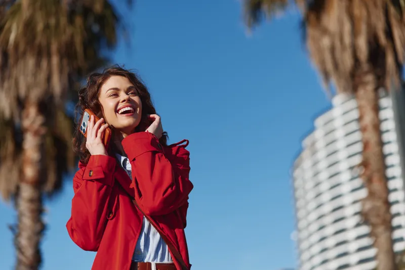 Young woman in a red jacket smiles while talking on a phone outdoors, with palm trees and a modern building in the background, capturing a bright candid moment.