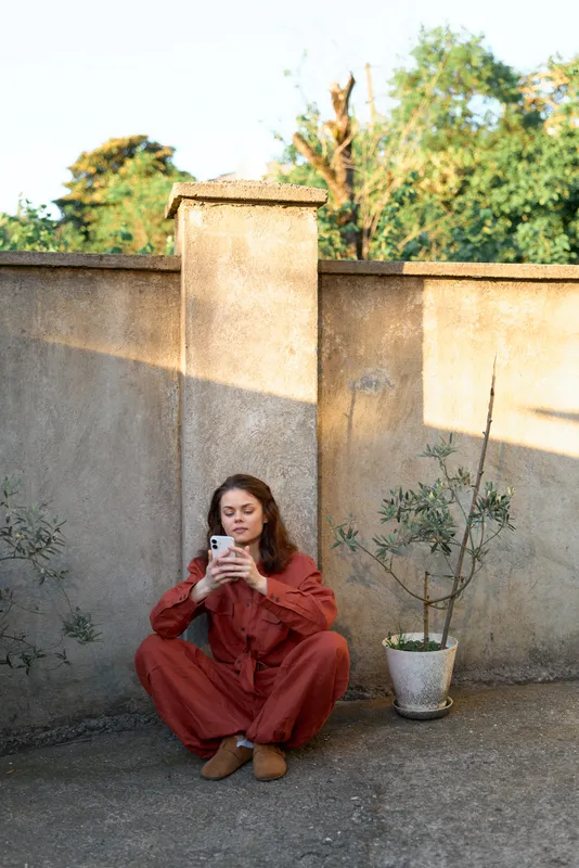 Woman outdoors, seated on the ground, wearing rust jumpsuit, focused on her phone, warm sunlight, calm wall backdrop, natural colors, candid lifestyle moment