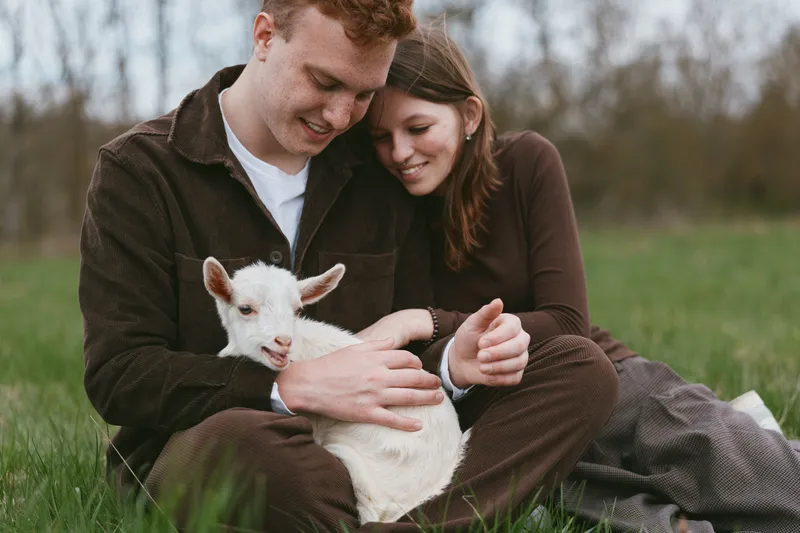 Young couple sitting on grass in casual clothes gently holding a white baby goat outdoors in early spring, sharing a tender and joyful moment together in nature
