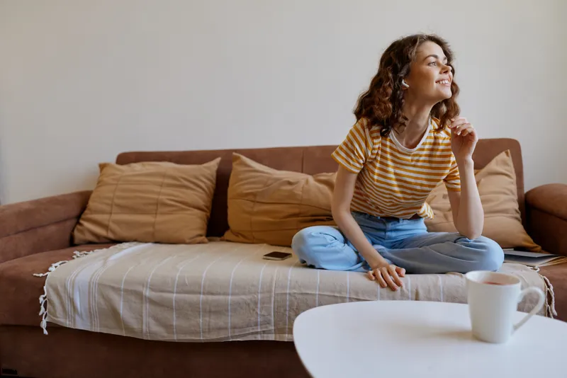 Smiling woman sitting on a couch in a cozy home, enjoying a relaxed lifestyle. Bright natural light fills the living room, cushions and table create a warm, comfortable scene.