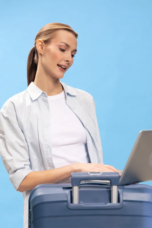 Young woman in casual clothes using laptop on suitcase with blue background. Travel concept, remote work and planning, lifestyle and modern technology in studio.