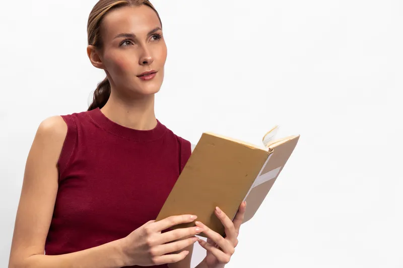 Young woman in red sleeveless top reading a book with focused expression against white background. Concept for education, knowledge, learning, and concentration.