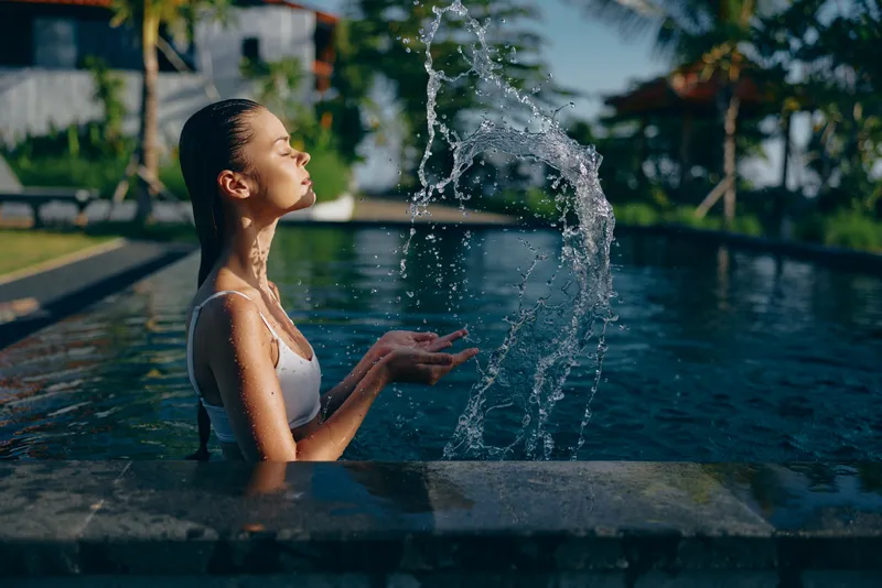 Young woman in white swimsuit enjoys relaxing time in outdoor pool playing with water splash under bright sunlight in tropical resort environment with palm trees background.