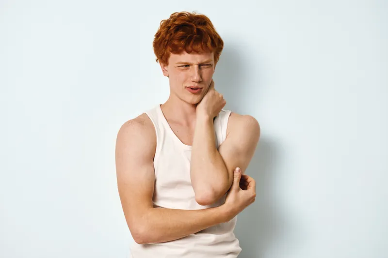 Young man with red hair wearing white tank top, showing pain and discomfort while touching his neck and elbow against white background, health and injury concept.