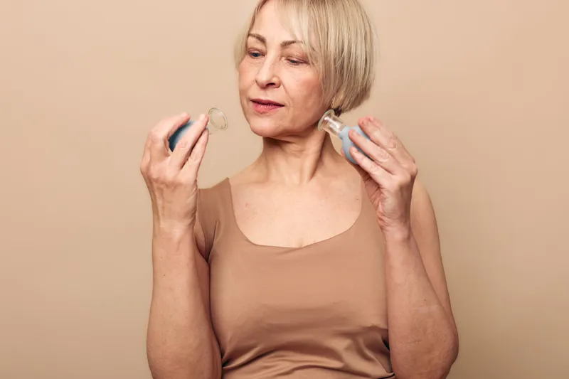 Mature woman with short blonde hair holds two hearing aids, examining them carefully against beige background. Concept of hearing health, aging, and assistive technology for seniors.