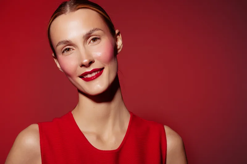 Smiling woman with red lipstick and red sleeveless top poses against red background. Close-up portrait with natural skin glow and confident expression. Concept of beauty and fashion.