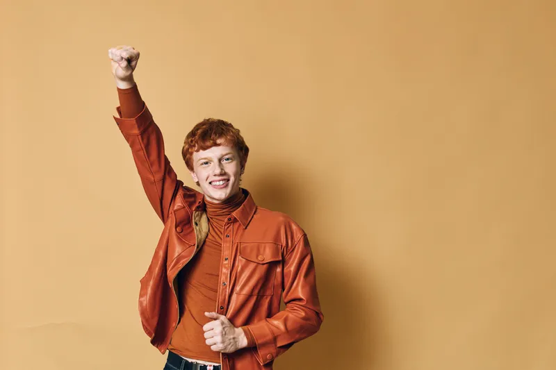 Young man with red hair wearing brown leather jacket and turtleneck smiling and raising fist on beige background for success and confidence concept.