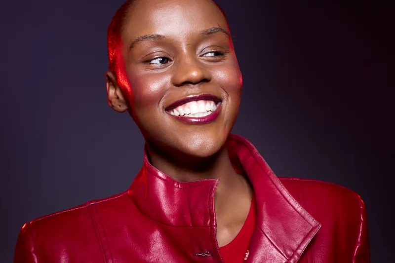 Smiling African woman with dark skin wearing red leather jacket looks away. Studio portrait with dramatic lighting and vibrant colors. Concept of fashion, confidence, and beauty.