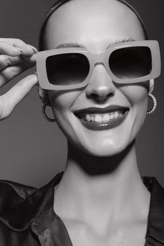 Smiling Caucasian woman with fair skin wearing large sunglasses and hoop earrings, dressed in a stylish blouse. Studio portrait in black and white, fashion and beauty concept.