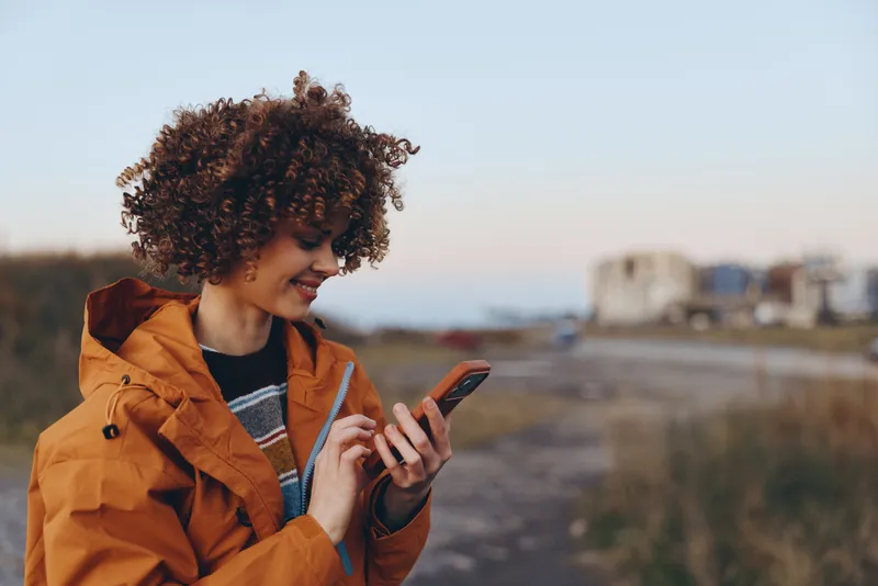 Woman outdoors, smiling while using smartphone, wearing orange jacket and curly hair, relaxed lifestyle moment by road, soft sunset light, urban background blur, distant vehicles