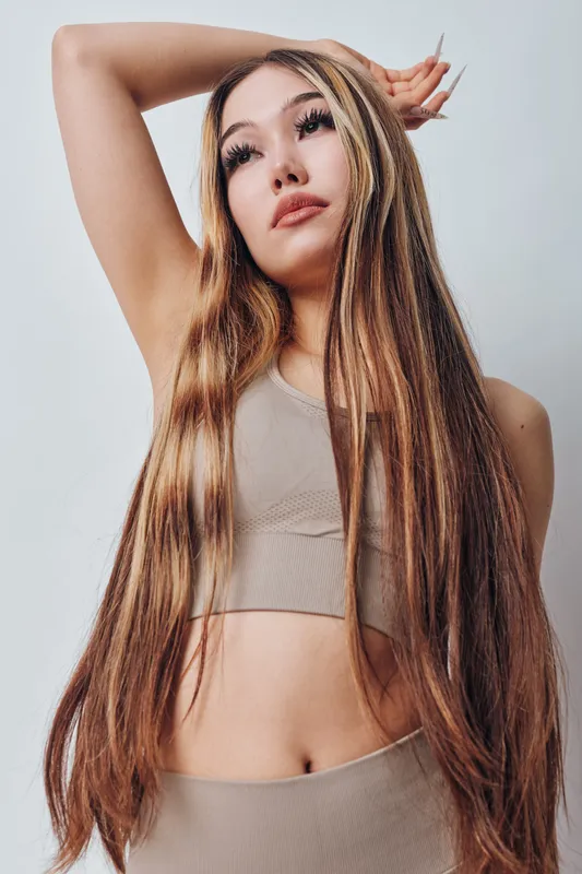 Young woman with long straight hair and natural makeup wearing beige crop top and pants posing with arm raised against light background for fashion and beauty concept.