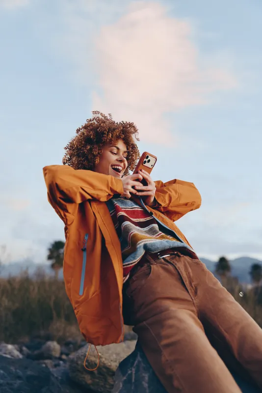 Smiling woman taking a selfie with a smartphone outdoors in a jacket, relaxed lifestyle in nature, soft clouds sky background, casual travel mood, self expression and joy, warm daylight