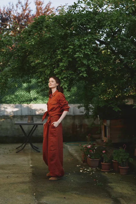 Woman in red jumpsuit stands in a garden, showcasing relaxed outdoor lifestyle, calm pose, soft natural light, potted plants and greenery framing her profile