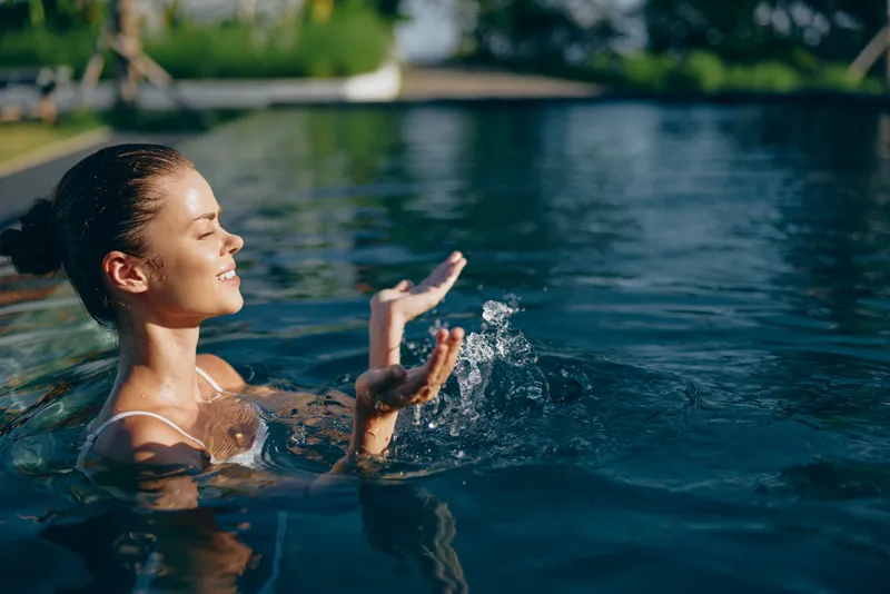 Relaxed woman enjoys water in a swimming pool, raising hands to splash as sunlight glints on blue water, embodying outdoor summer leisure and calm mood