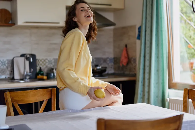 Cheerful woman indoors, relaxed lifestyle moment, sitting on a kitchen table, lemon in hand, sunlit room, warm colors, candid smile, casual home setting, everyday joy, natural mood