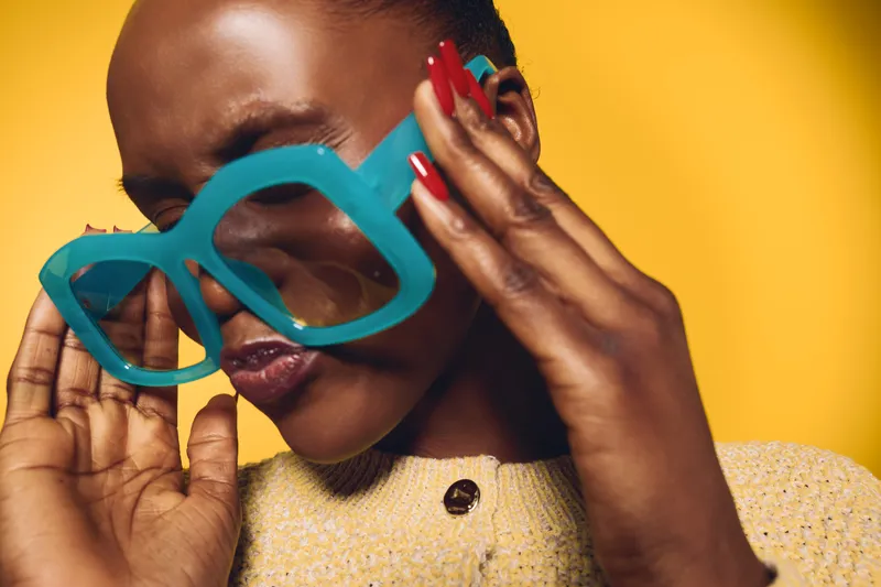 Close-up of a dark skinned African woman wearing large blue sunglasses and a yellow sweater. She touches her face with red manicured nails against a bright yellow background.