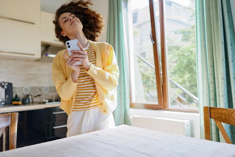 Young woman in a sunlit kitchen wears a striped tee and yellow cardigan, holds a smartphone, head tilted back, enjoying a carefree moment by the window, showing relaxed curiosity and warm mood.