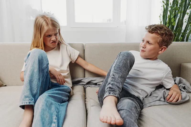 Unhappy girl and smiling boy sitting on couch in casual clothes in bright room, showing sibling emotions and interaction at home with natural light.