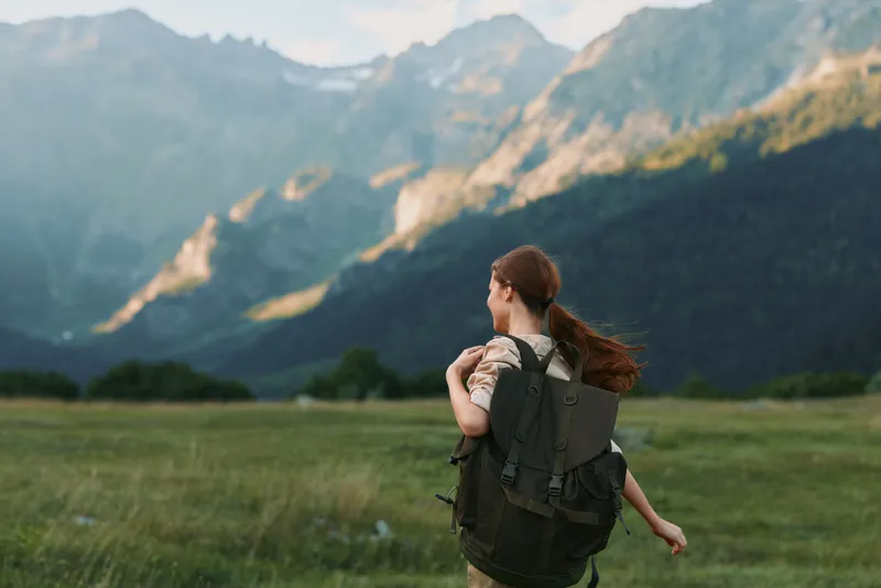 A young woman with a backpack walks through a sunlit grassy field toward distant mountains, exuding adventure, freedom, and a hopeful mood as a light breeze rustles her hair