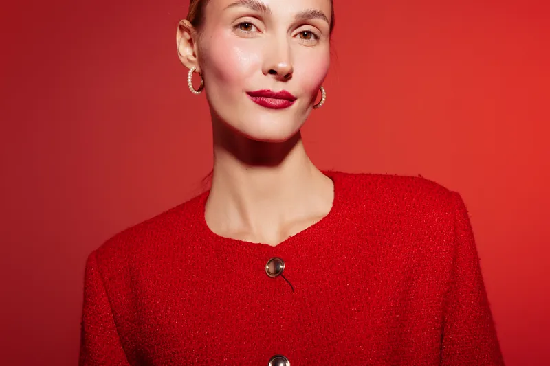 Confident woman in red jacket poses against red background. She wears hoop earrings and red lipstick, creating a bold and stylish look. Concept for fashion, beauty, and elegance.