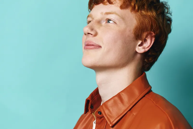 Young man with red hair wearing a brown leather jacket smiling and looking up against a blue background. Casual portrait with natural expression and clean studio lighting.