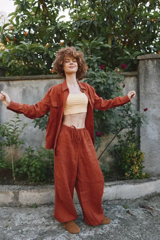 Young woman with curly hair dances in a rust orange tracksuit in a garden, arms wide, radiating confidence and carefree energy