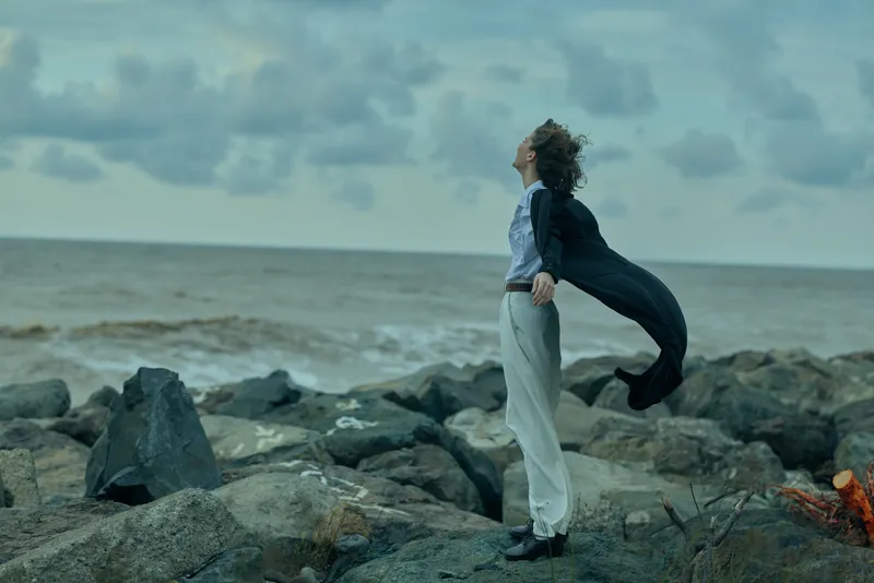 Figure stands on rocky shoreline with cape flowing in wind, eyes toward the horizon, creating a moody coastal moment that conveys freedom, resilience, and reflection against a stormy sea.