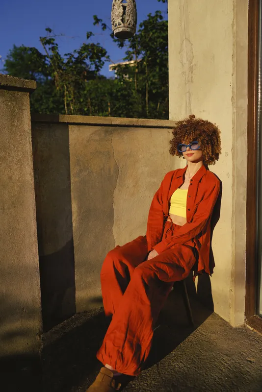 Stylish one person woman with curly hair sits on a sunlit terrace in a bright orange suit and yellow top, creating a confident fashion lifestyle portrait for a relaxed editorial mood