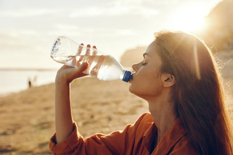 Woman drinking water outdoors in sunlight on a beach, hydration and wellness focus, healthy adult female in profile with plastic bottle, closeup face and calm expression, summer travel nature