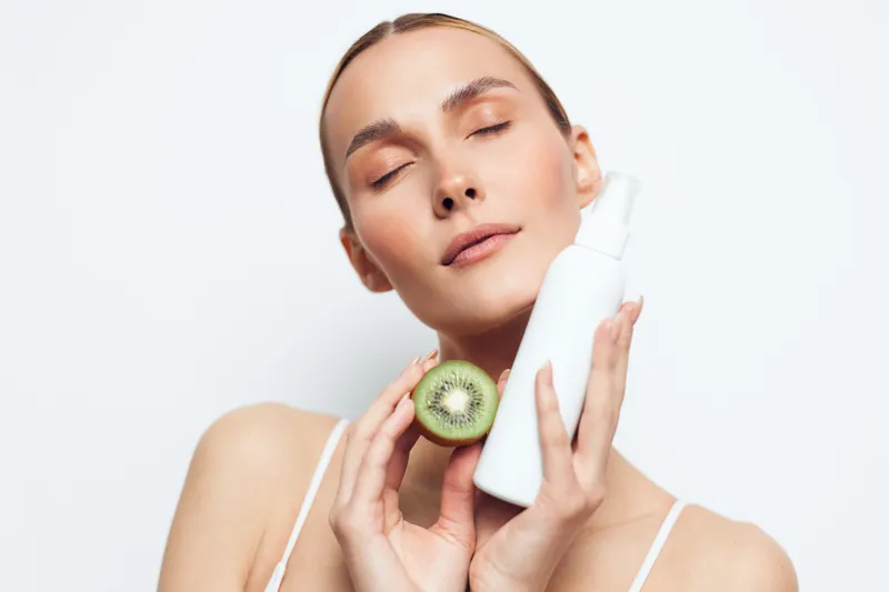 Young woman with closed eyes holding a white skincare bottle and a fresh kiwi fruit, isolated on white background. Concept of natural beauty and facial care.