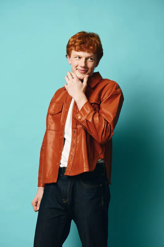 Young man with red hair wearing brown leather jacket and white t shirt posing with hand on chin against blue background, casual fashion and confident expression.