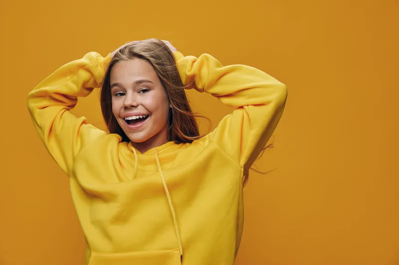 Happy girl smiling with hands behind head wearing yellow hoodie against orange background, casual portrait of cheerful teenager for lifestyle and youth fashion concept.