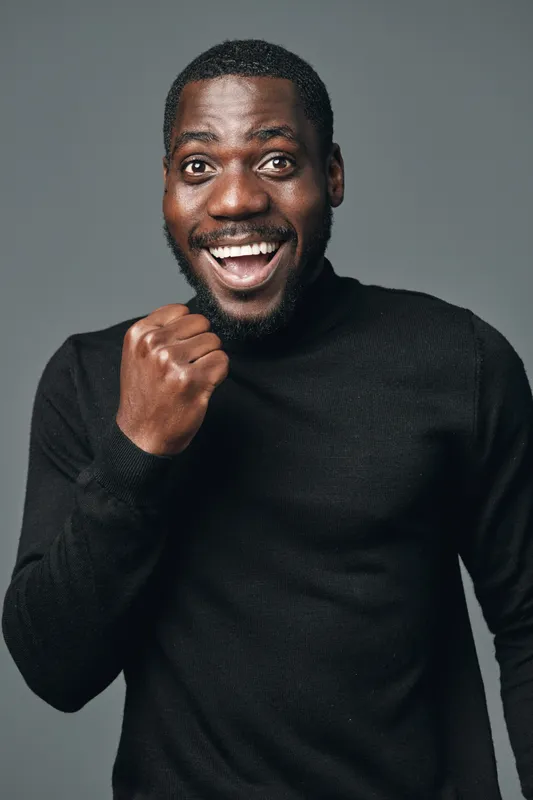 African man with dark skin wearing black sweater showing excitement and joy with clenched fist and wide smile in creative conceptual visual style on gray background.
