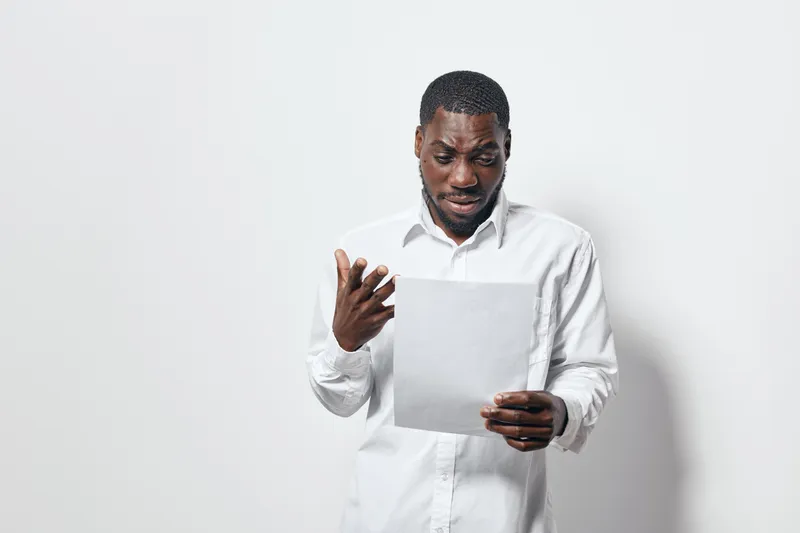African man with dark skin reading document with confused expression in white shirt on white background. Male person holding paper and looking surprised or worried indoors.