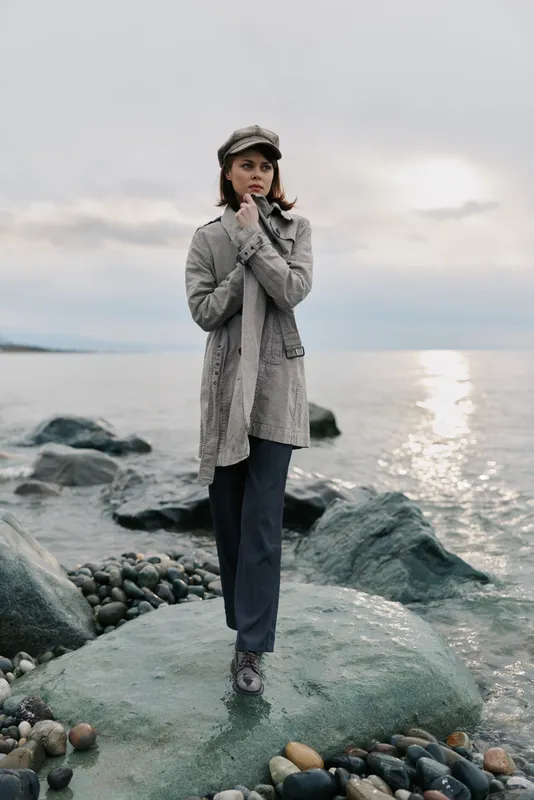 Young woman in grey coat and cap stands on rock by the sea on cloudy day. She looks thoughtful and warm, surrounded by stones and water reflecting soft light. Concept of autumn fashion.