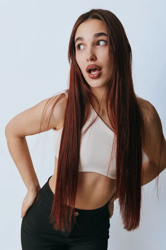 Young woman with long straight hair wearing a sleeveless top and black pants posing with hands on hips against white background looking to the side with open mouth expression.