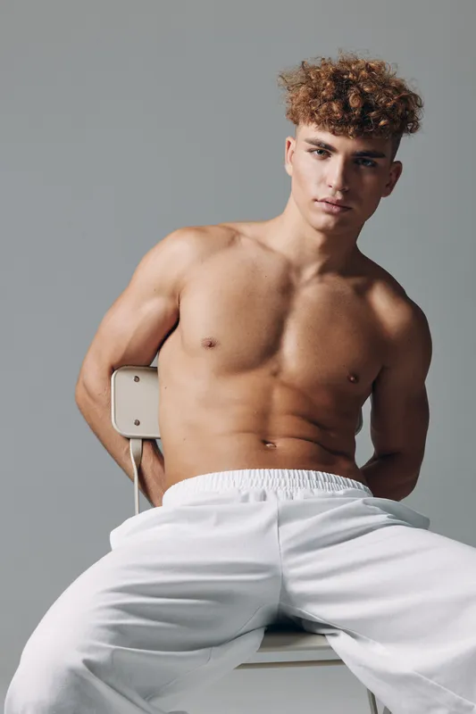 Young man with curly hair sitting shirtless on a chair wearing white pants against plain background. Posed confident, fitness, casual and indoors studio with soft lighting.