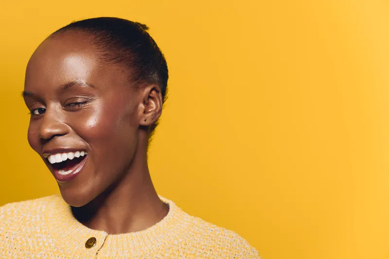 Happy smiling African woman with dark skin winks and laughs wearing yellow sweater on bright yellow background. Concept of joy, beauty, positive emotions, and confident female portrait.