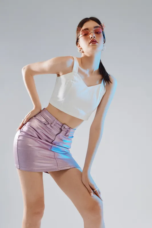 Young woman in a white crop top and metallic purple skirt poses confidently against a neutral background. Concept for woman beauty fashion and modern style.