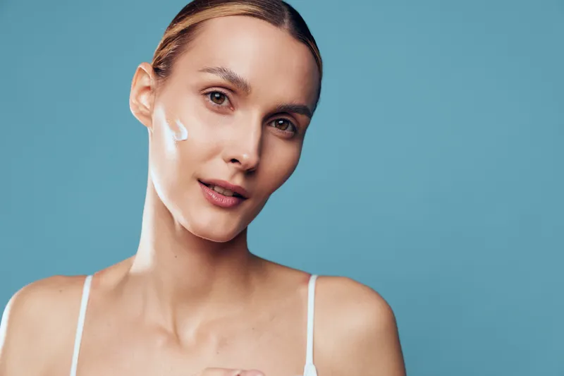 Young woman with light skin applies facial cream to her cheek in a bright studio, blue background, calm mood, self care ritual, skincare routine, minimal makeup, focused expression.