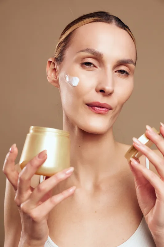 Young woman with blonde hair applies cream on her cheek while holding an open jar in a skincare routine against a neutral background with a calm expression.